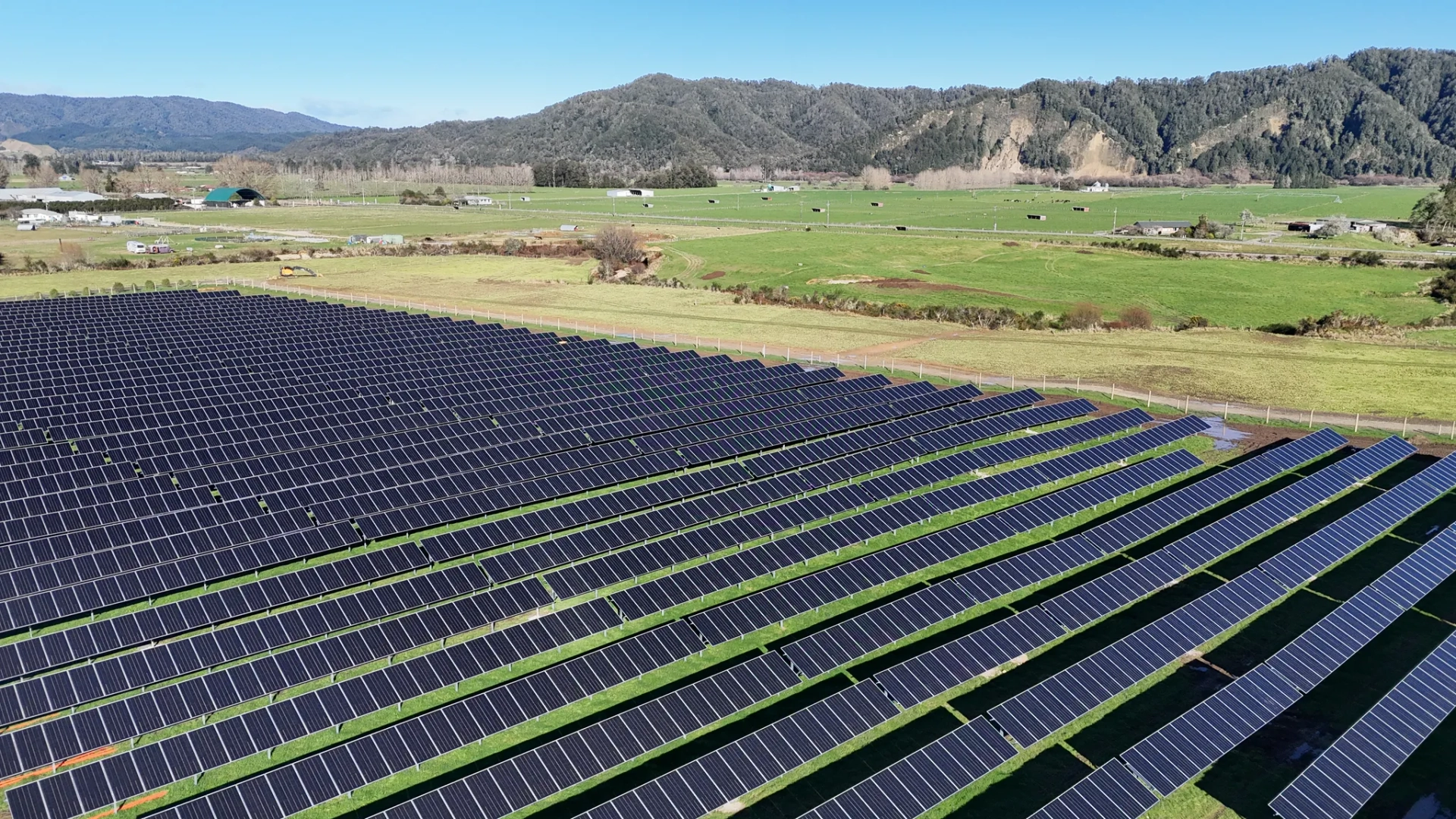 Aerial view of solar panel rows at Reefton Solar Farm with forested hills behind