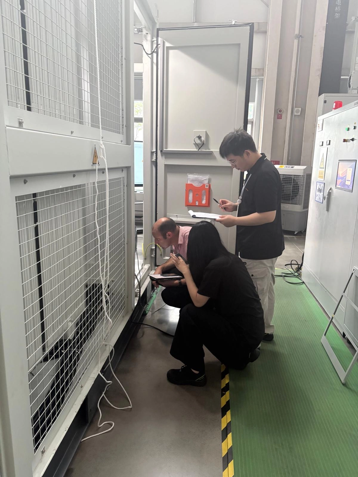 Three people inspecting electrical equipment inside a factory